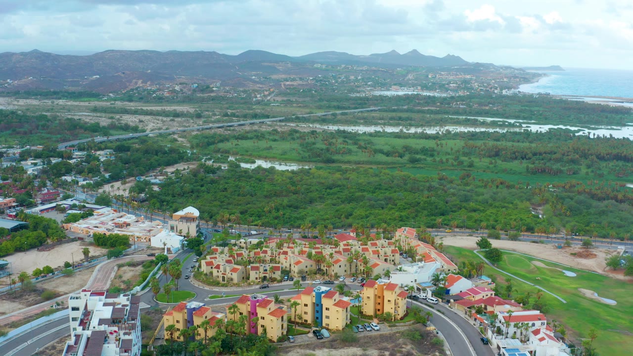 San Jose Del Cabo aerial frame with yellow housing and a curved road along the bottom edge, green land and water across the center, hills on the horizon, coastline at right, and no visible people.