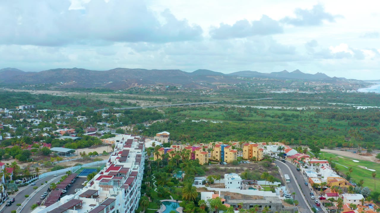 Wide aerial frame over San Jose Del Cabo showing foreground apartment blocks and roads, green lowland, distant hills, coastal water at right, and no visible people.