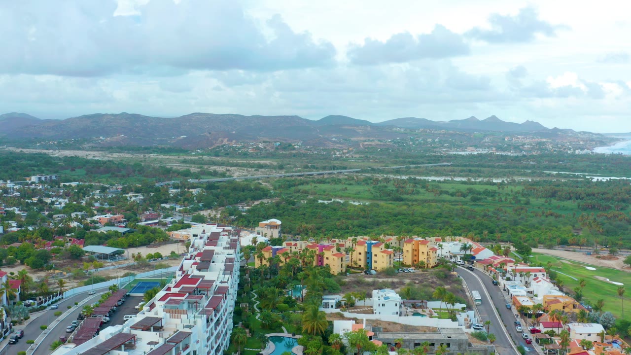 Drone view across colorful buildings, roadway, green terrain, distant hills, and water.