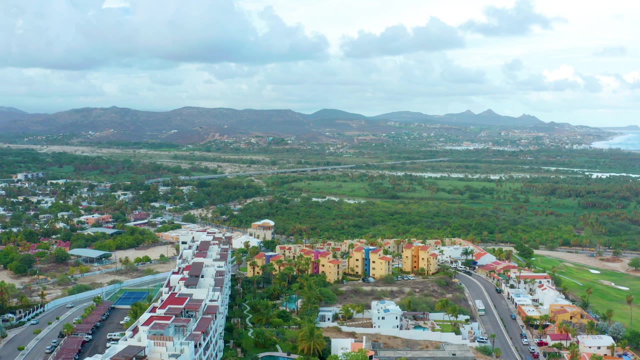 High drone frame over San Jose Del Cabo showing a larger spread of foreground housing, green terrain through the center, hills in the background, coastal water on the right edge, and no visible people.