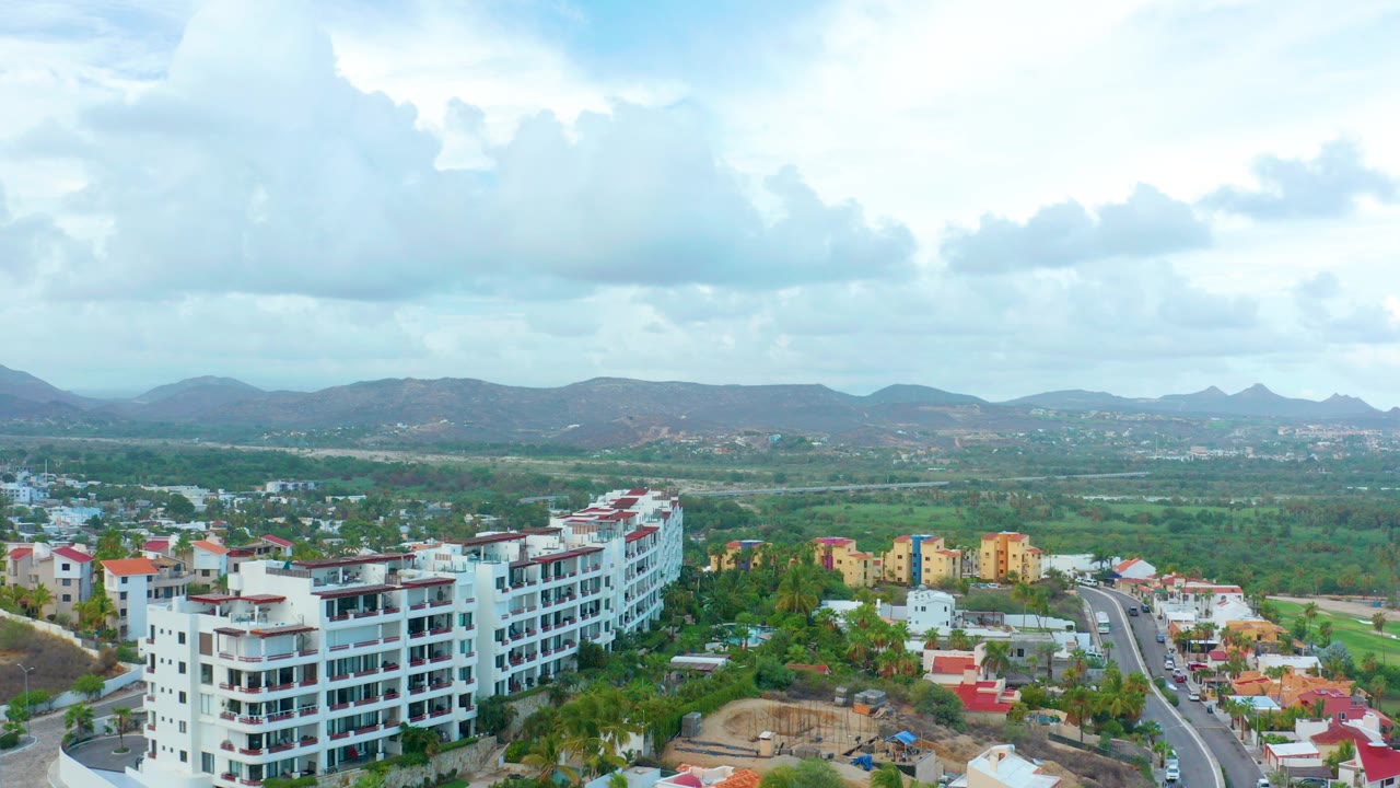 High aerial frame over San Jose del Cabo showing white apartment blocks at left, smaller homes below, green valley, distant mountains, partly cloudy sky, and no visible people.