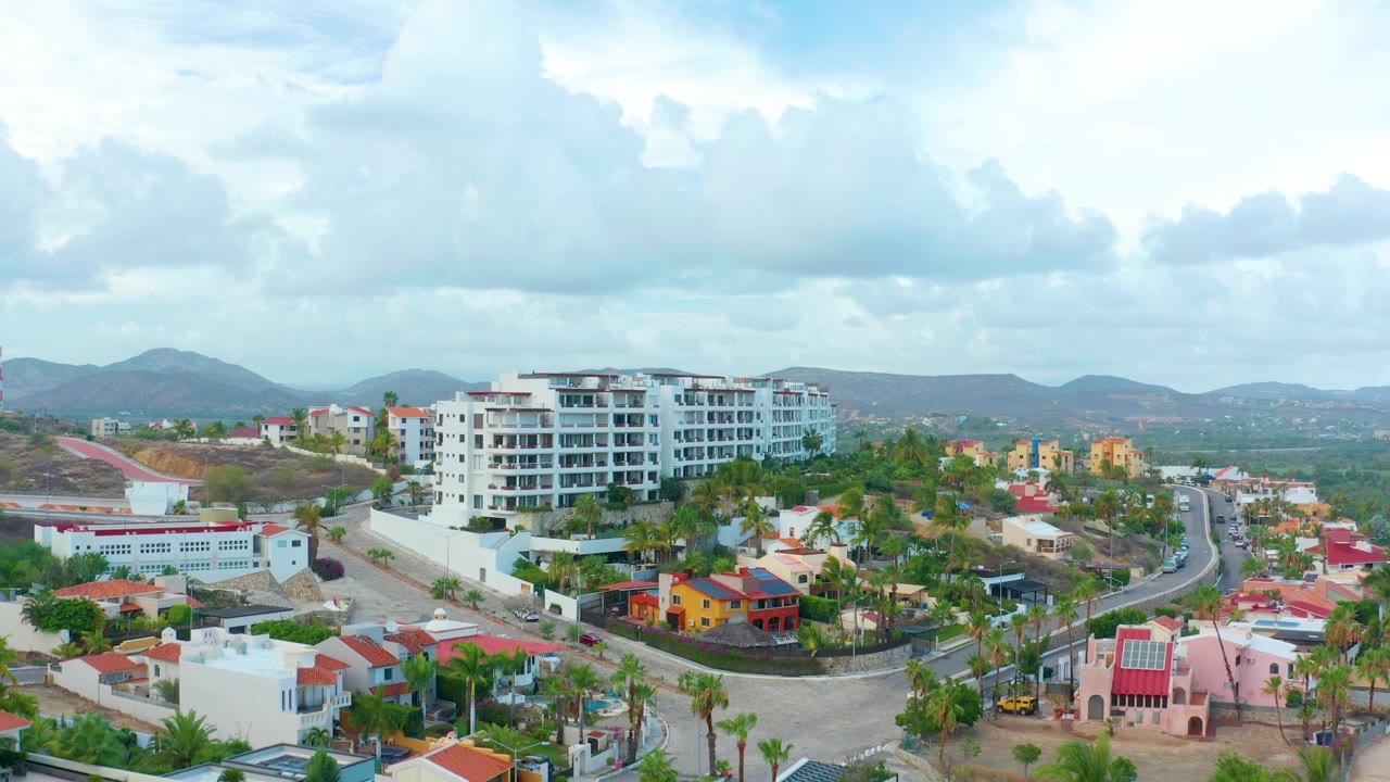 Opening aerial frame over San Jose del Cabo with white apartment buildings near center, red-roofed homes in the foreground, distant mountains, cloudy sky, and no visible people.