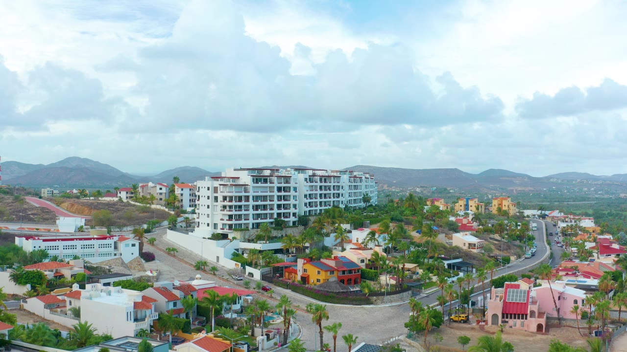 Aerial neighborhood view with apartment blocks, colorful houses, winding road, palms, hills, and clouds.