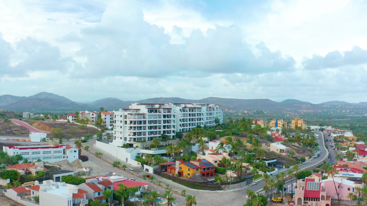 San Jose del Cabo aerial frame looking over red-roofed homes and palms toward a white residential building, green valley, low mountains, cloud-filled sky, and no visible people.