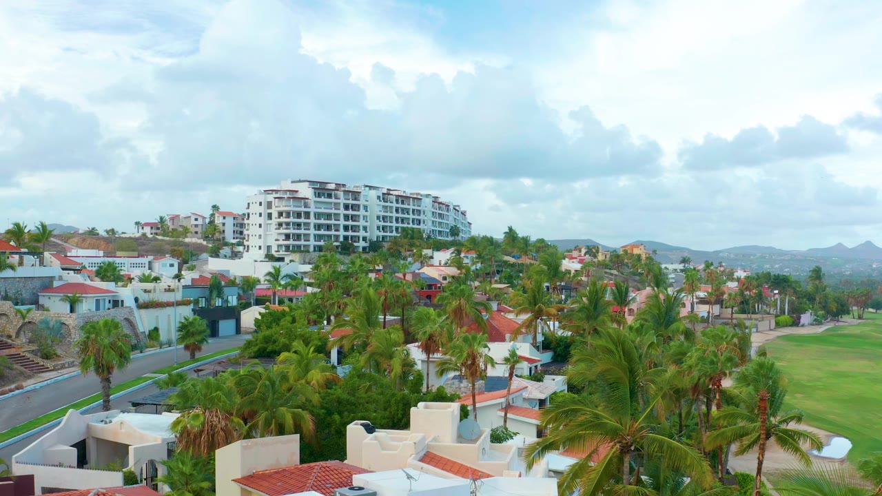 Opening frame of San Jose Del Cabo, Baja California Sur, with palms and white homes in the foreground, a green lawn at right, a white multi-story building beyond, and no visible people.