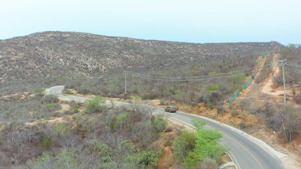 Aerial road view in La Playita Los Cabos, Mexico, with the vehicle slightly farther along the curve, scrub-covered slopes, utility lines, exposed dirt, and no visible people.