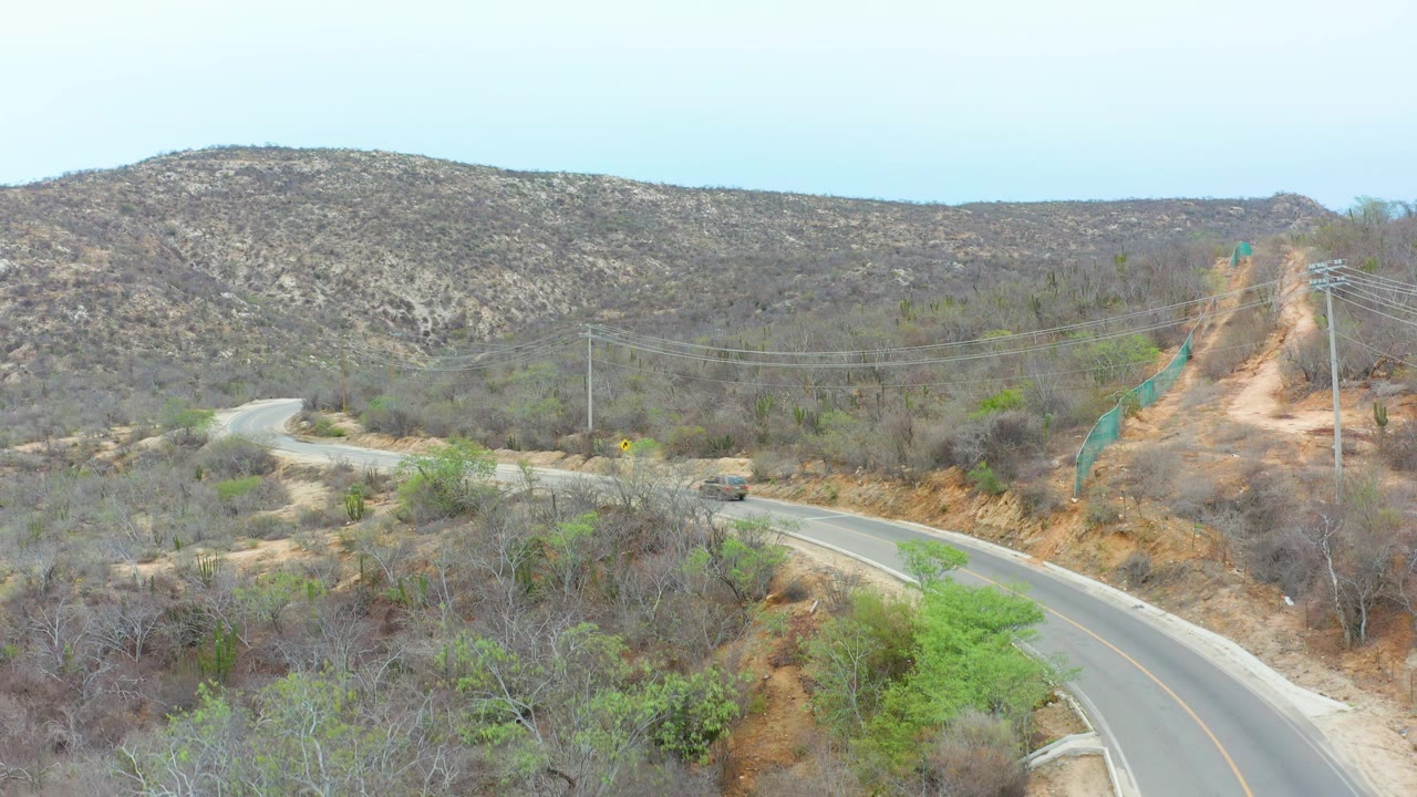 Elevated frame of the La Playita Los Cabos road as the vehicle nears the bend, with brown brush, green shrubs, utility poles, pale sky, and no visible people.