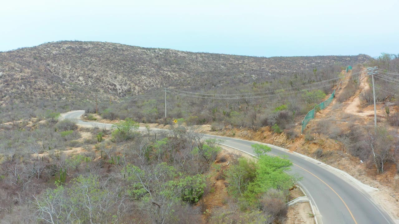 Aerial view over a winding paved road in La Playita Los Cabos, Mexico, with the vehicle advanced along the left road section, dry vegetation, power lines, and no visible people.