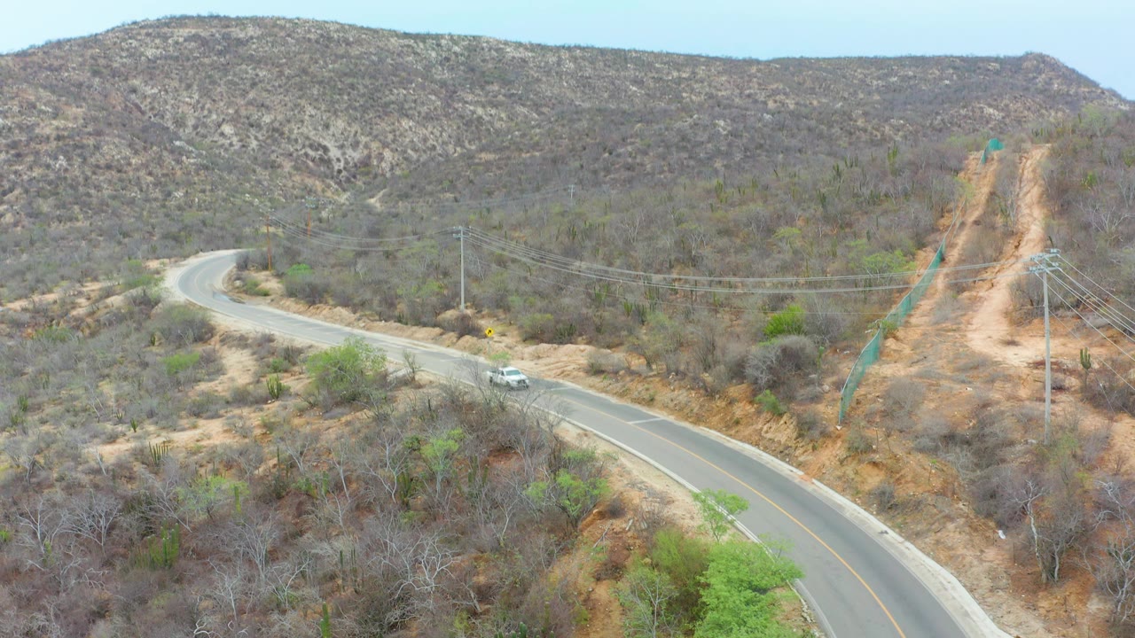 Aerial view in La Playita Los Cabos with the white car slightly farther along the winding road, scrubby hills behind it, utility lines crossing the scene, and no visible people.