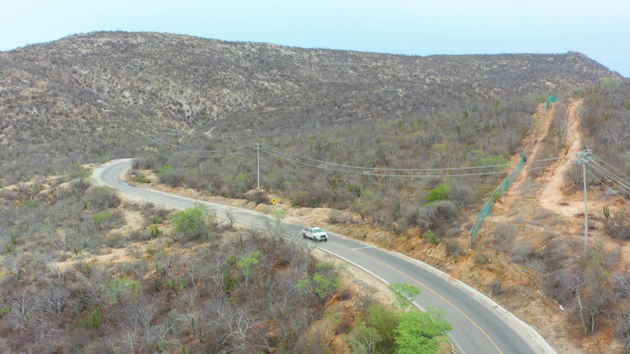 La Playita Los Cabos road frame showing the white car approaching the lower curve, a tan dirt cut on the hillside, utility poles and wires, dry brush, and no visible people.