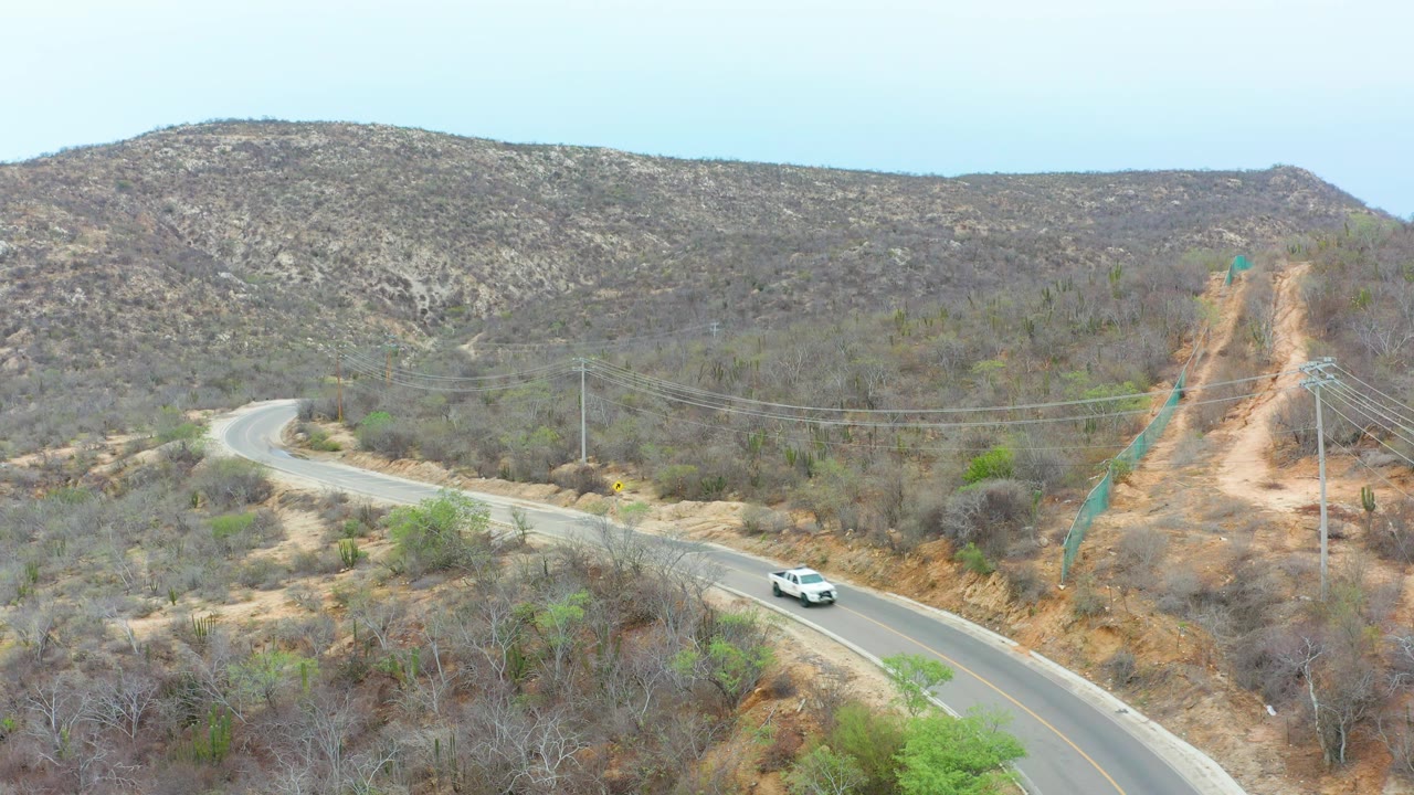 Aerial still from La Playita Los Cabos with the white car nearing the foreground curve, the road bending left through dry vegetation, hills across the background, and no visible people.