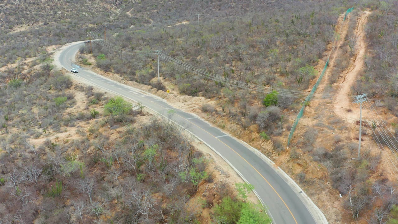 Aerial view in La Playita Los Cabos with the white car slightly farther along the road curve, dry brush on both sides, a dirt utility track, and no visible people.