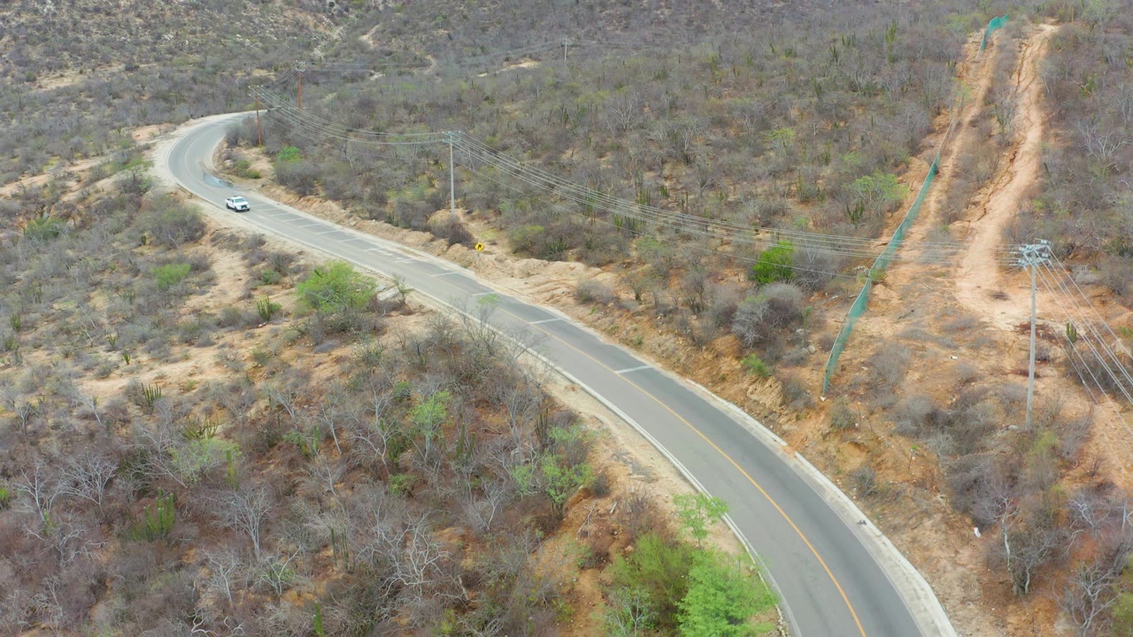 In La Playita Los Cabos, the white car sits near the left side of the winding road while dry gray-brown hills, scattered green shrubs, power lines, and no visible people fill the frame.