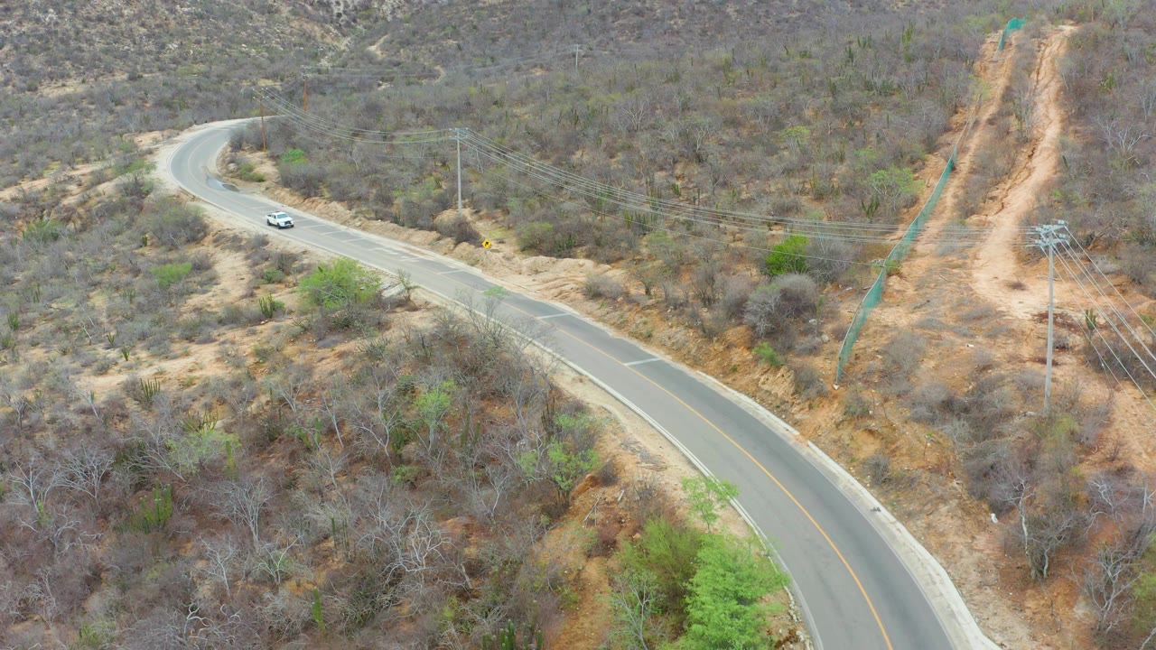 Aerial frame from La Playita Los Cabos showing the white car on the upper road segment, a sweeping paved curve below, a dirt cut up the hill, and no visible people.