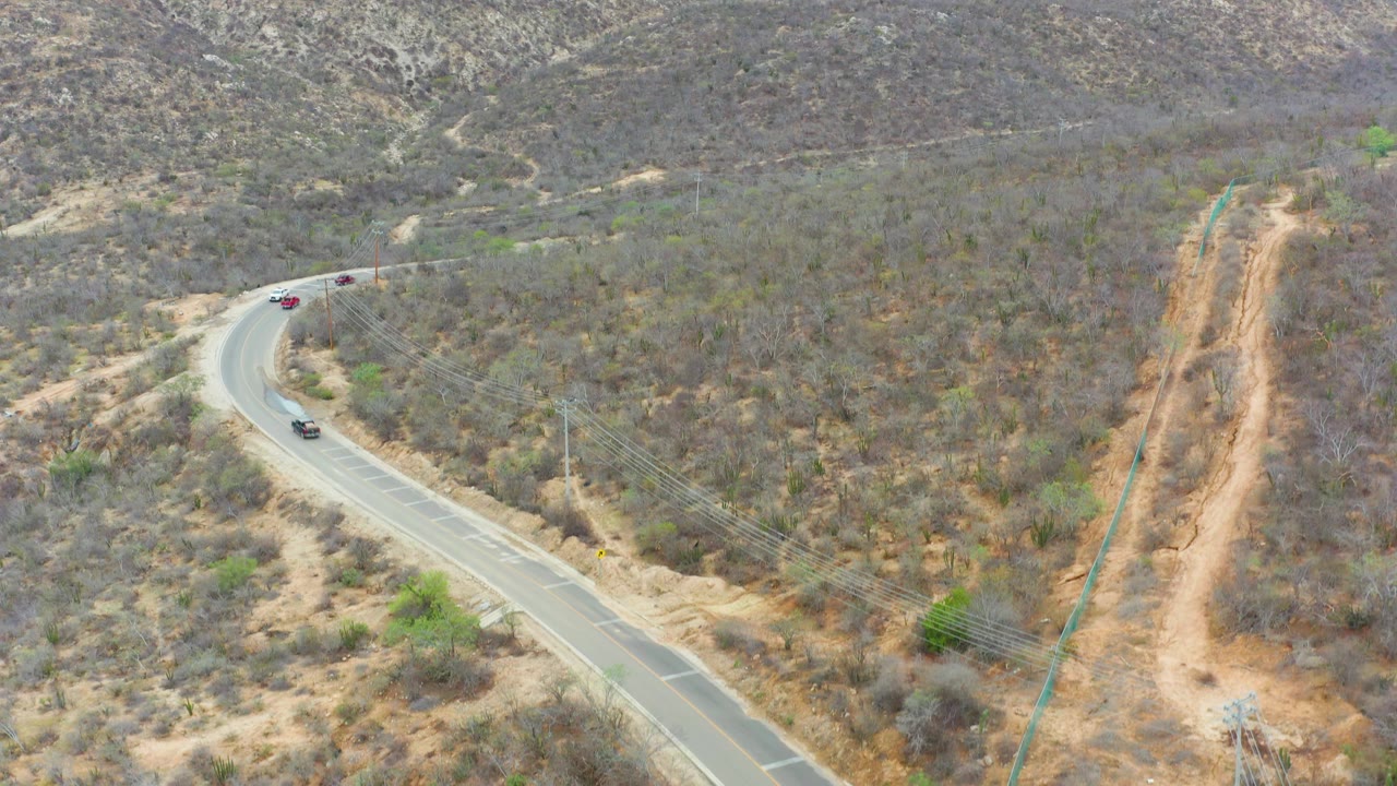 Aerial view in La Playita Los Cabos, Mexico, with vehicles spaced along the road curve, a tan dirt corridor beside utility lines, dry brush, and no visible people.