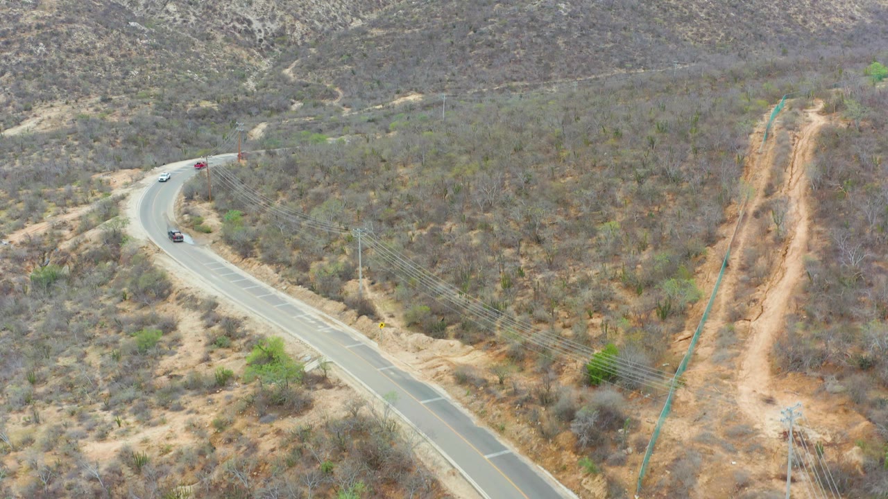Aerial frame from La Playita Los Cabos, Mexico, showing cars on a pale road, a steep dirt cut with power infrastructure to the right, scrub vegetation, and no visible people.