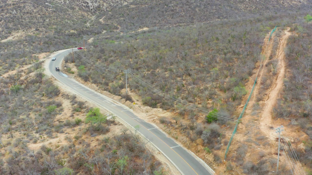 La Playita Los Cabos aerial frame with a car near the upper road bend, the road sweeping into the lower frame, dry brush on both sides, utility lines, and no visible people.