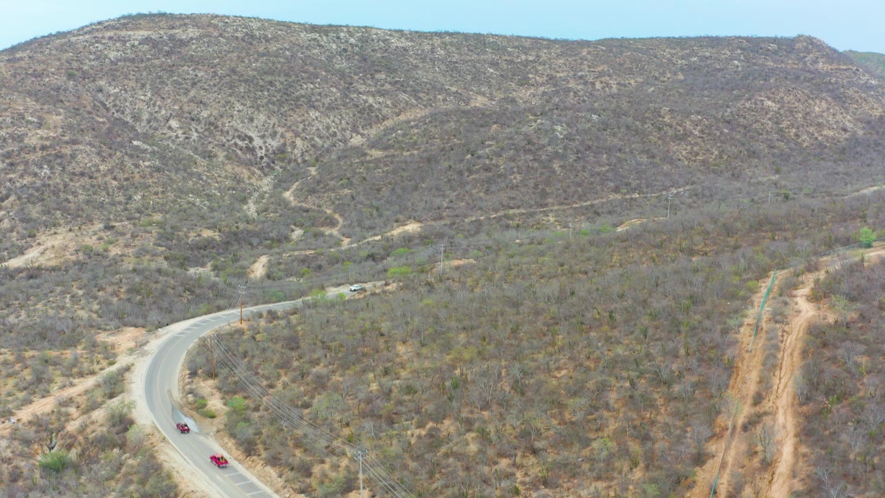 Aerial view of two small vehicles entering a curved road through dry scrub hills in La Playita Los Cabos, Mexico, with no visible people.