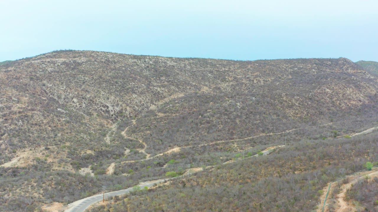 Aerial frame of scrub-covered hills in La Playita Los Cabos, Mexico, with the paved road curving along the lower edge and no visible people.