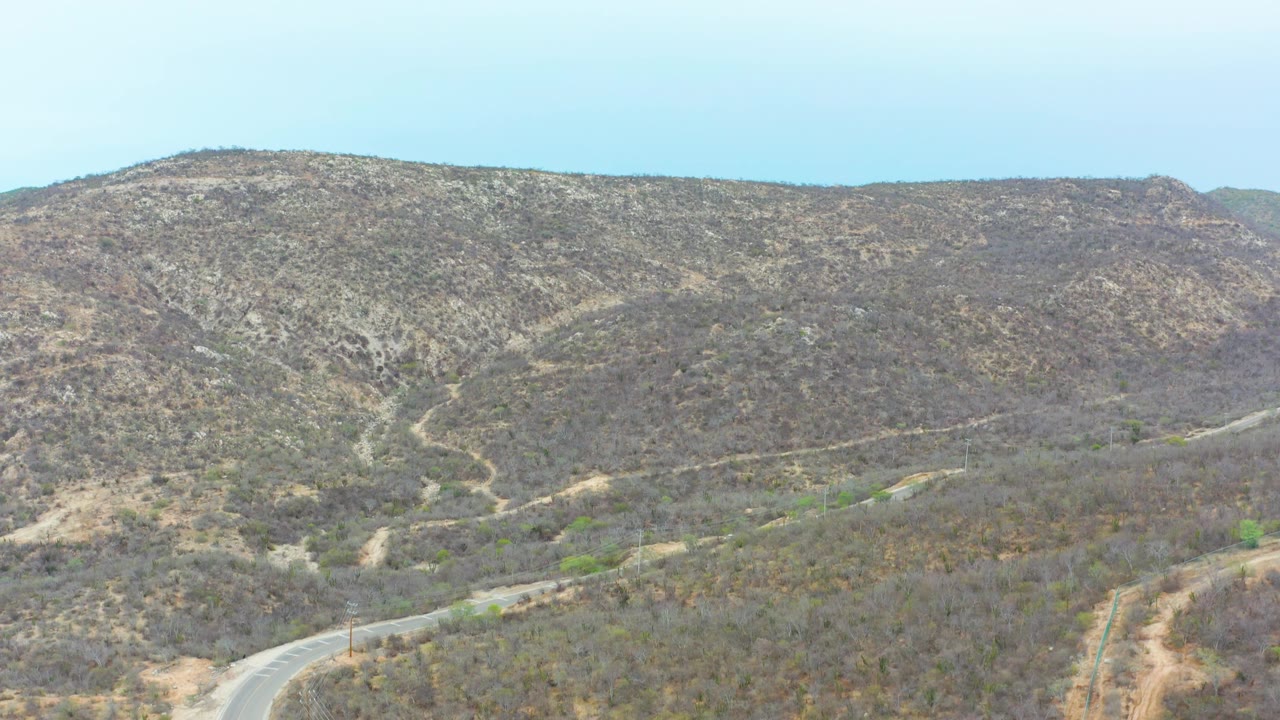 Wide aerial view in La Playita Los Cabos, Mexico, showing arid hills, pale dirt tracks, and a road bend near the lower frame with no visible people.
