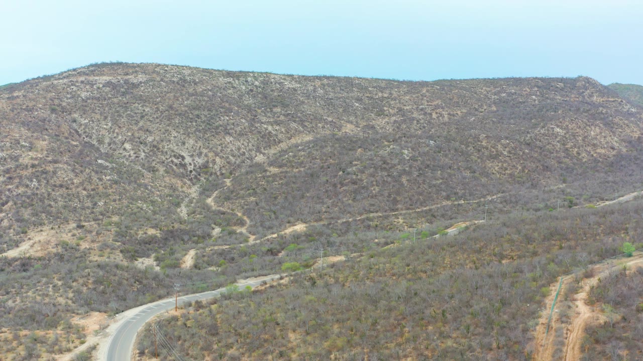 Aerial view of the La Playita Los Cabos hillside, with sparse brush across the slopes and the winding road visible at the lower left, with no visible people.