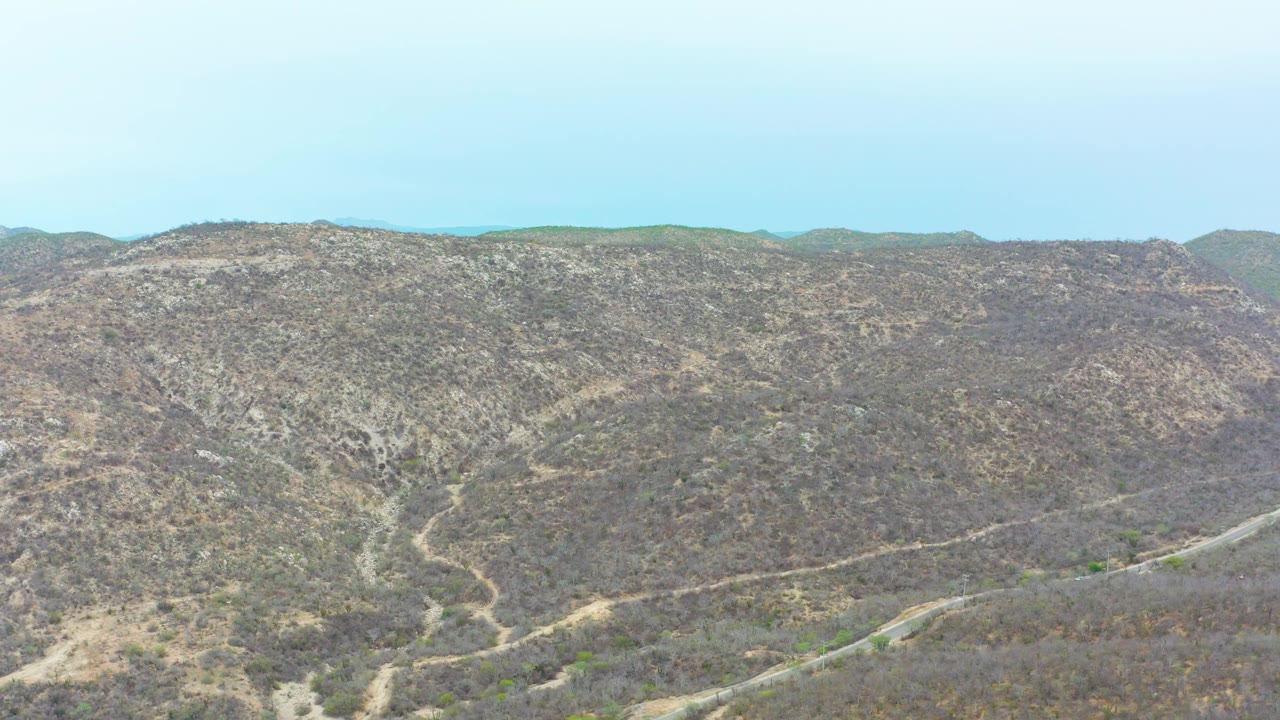 Wide elevated frame over La Playita Los Cabos arid hills, with a central slope of mottled scrub, thin dirt paths, pale sky, and no visible people.