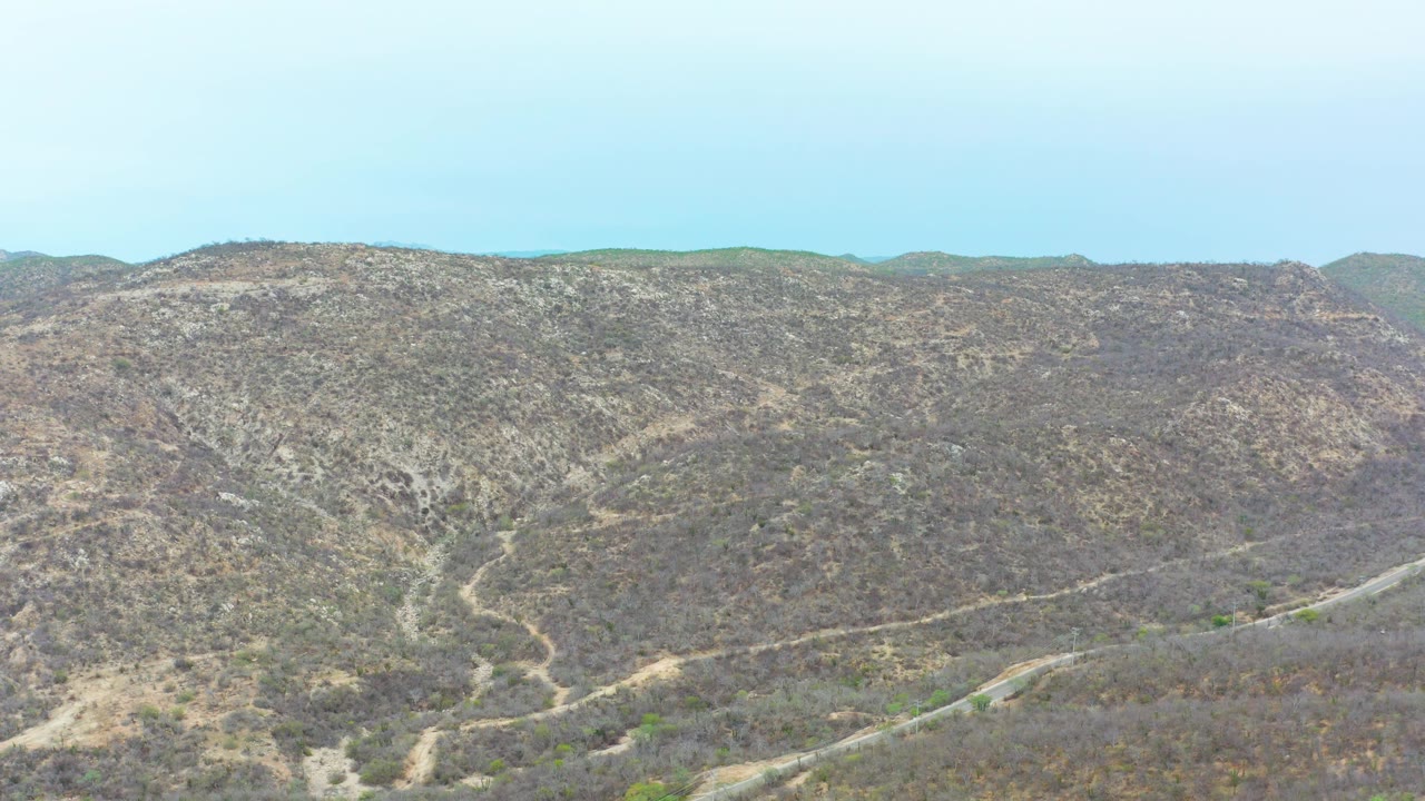 Mid-clip view of the La Playita Los Cabos hillside, where a light dirt path bends through gray-brown brush below a low distant horizon, with no visible people.