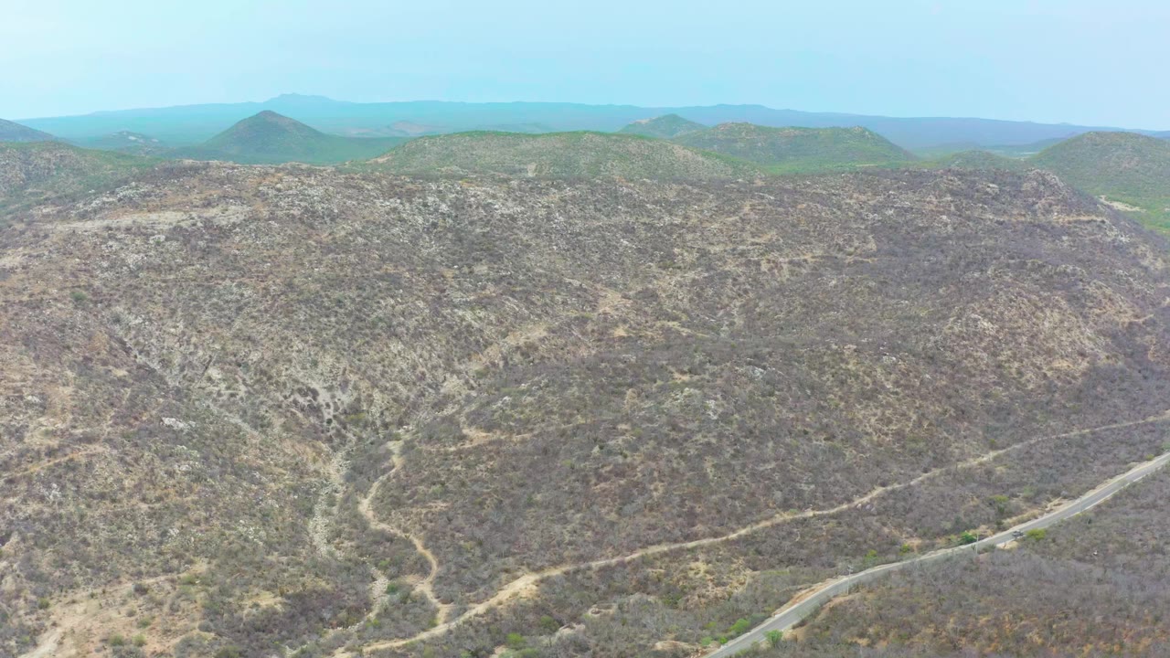 Wide aerial view in La Playita Los Cabos showing rocky arid hills, sparse scrub, a curving road near the lower right edge, distant rounded hills, and no visible people.