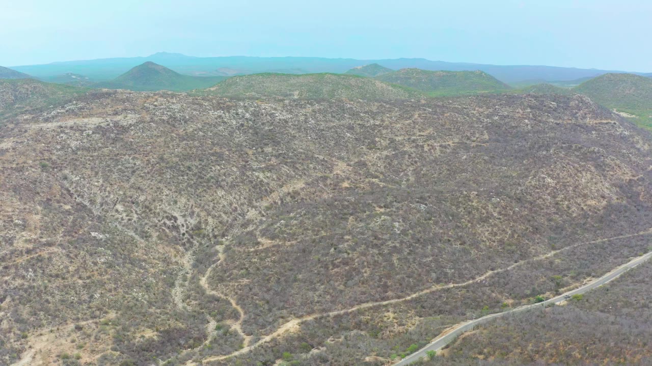 La Playita Los Cabos hillside frame with beige dirt tracks cutting through rocky scrub, the paved road visible along the lower right, layered hills on the horizon, and no visible people.