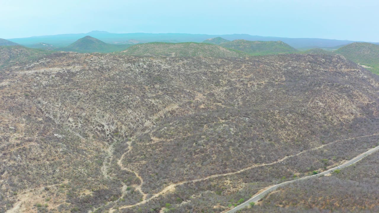 Wide elevated frame over La Playita Los Cabos with mottled brown hills, a winding road entering from the lower right, small dirt paths on the slopes, hazy background hills, and no visible people.