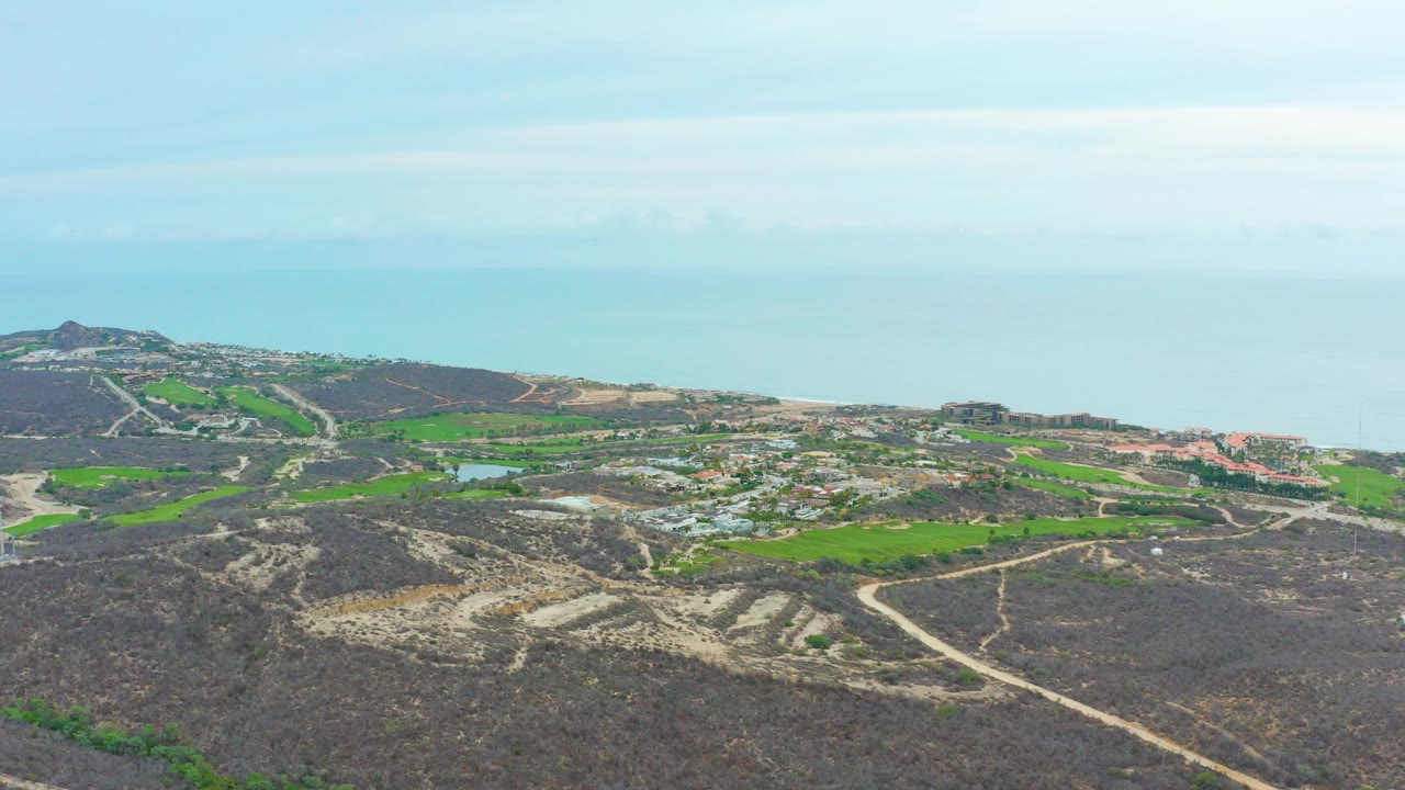 Opening frame of La Playita Los Cabos from above, showing dark rocky foreground, bright green field patches, winding dirt roads, scattered buildings, distant ocean, and no visible people.