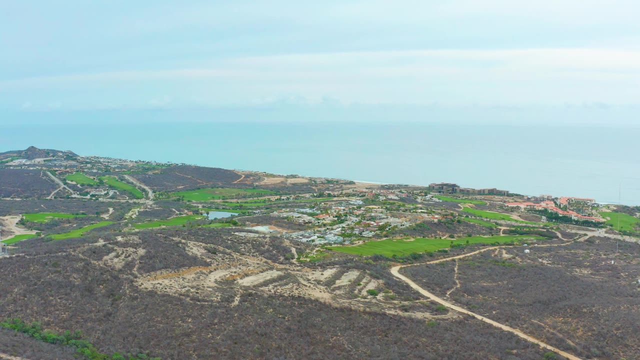 At 0:01, the La Playita Los Cabos aerial view holds on rocky brown terrain, green cultivated areas, pale roads, clustered structures, blue ocean horizon, and no visible people.