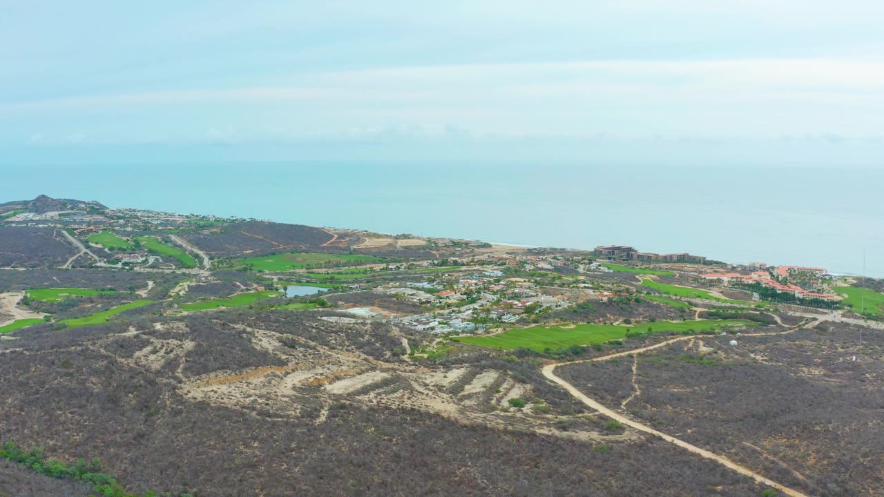 Wide coastal aerial with dark terrain, curving roads, green fields, buildings, and blue water.