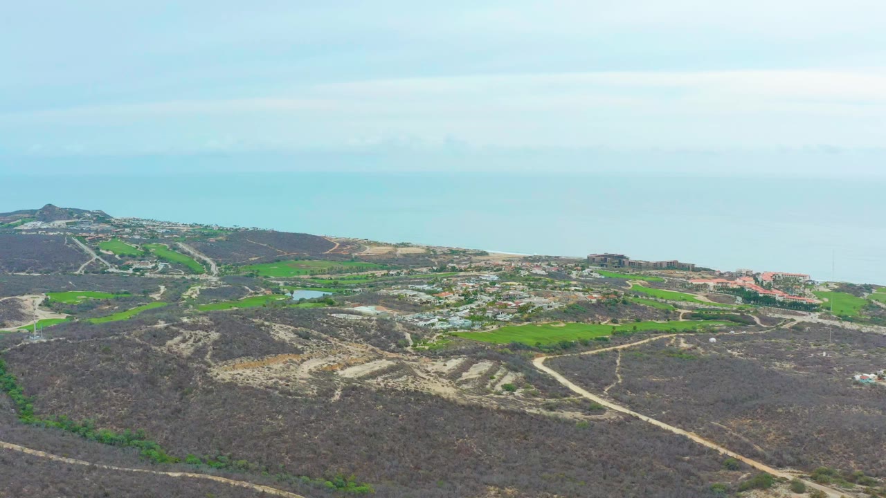 Wide aerial frame over La Playita Los Cabos, Mexico, showing dry hills crossed by pale dirt roads, green field patches, scattered buildings, ocean horizon, and no visible people.
