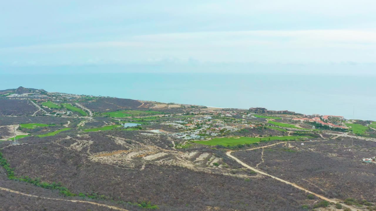 Aerial view in La Playita Los Cabos with winding roads cutting through dark scrub hills, brighter green fields near the center, buildings near the coast, and no visible people.
