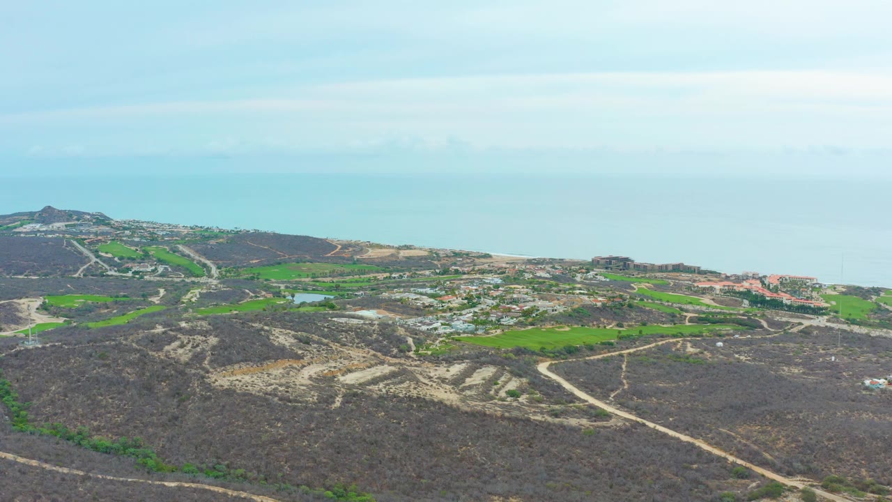 La Playita Los Cabos aerial landscape showing a pale road network over dry hilly ground, green agricultural-looking patches, clustered structures, blue ocean, and no visible people.