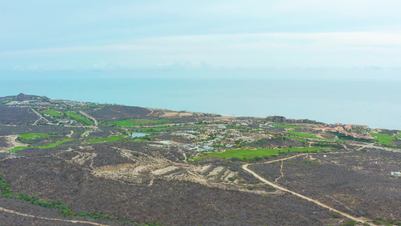 Coastal hillside aerial with dirt roads, green fields, settlement, and ocean under hazy sky.