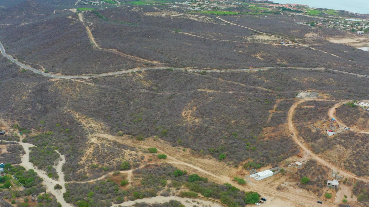 Aerial view over La Playita Los Cabos showing dry scrub hills, intersecting dirt roads, a few small buildings, distant developed land, and no visible people.