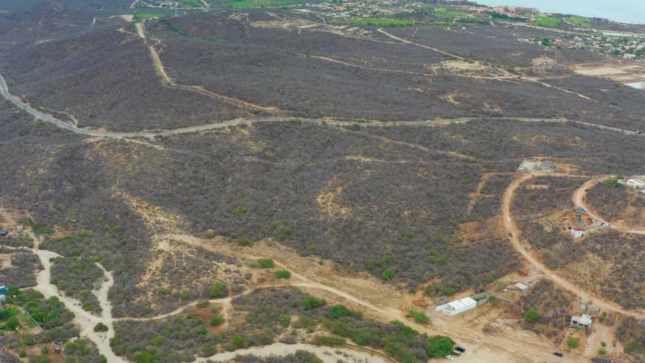 Aerial frame in La Playita Los Cabos with pale dirt tracks cutting through brown scrub hills, small roadside structures near the lower edge, distant roads, and no visible people.