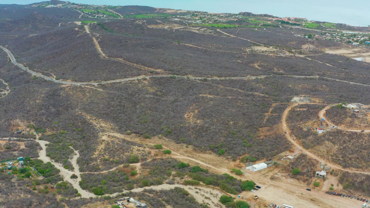 Wide drone frame over La Playita Los Cabos showing arid hills, pale dirt tracks crossing the slopes, a small structure to the right, distant roads on the ridge, and no visible people.