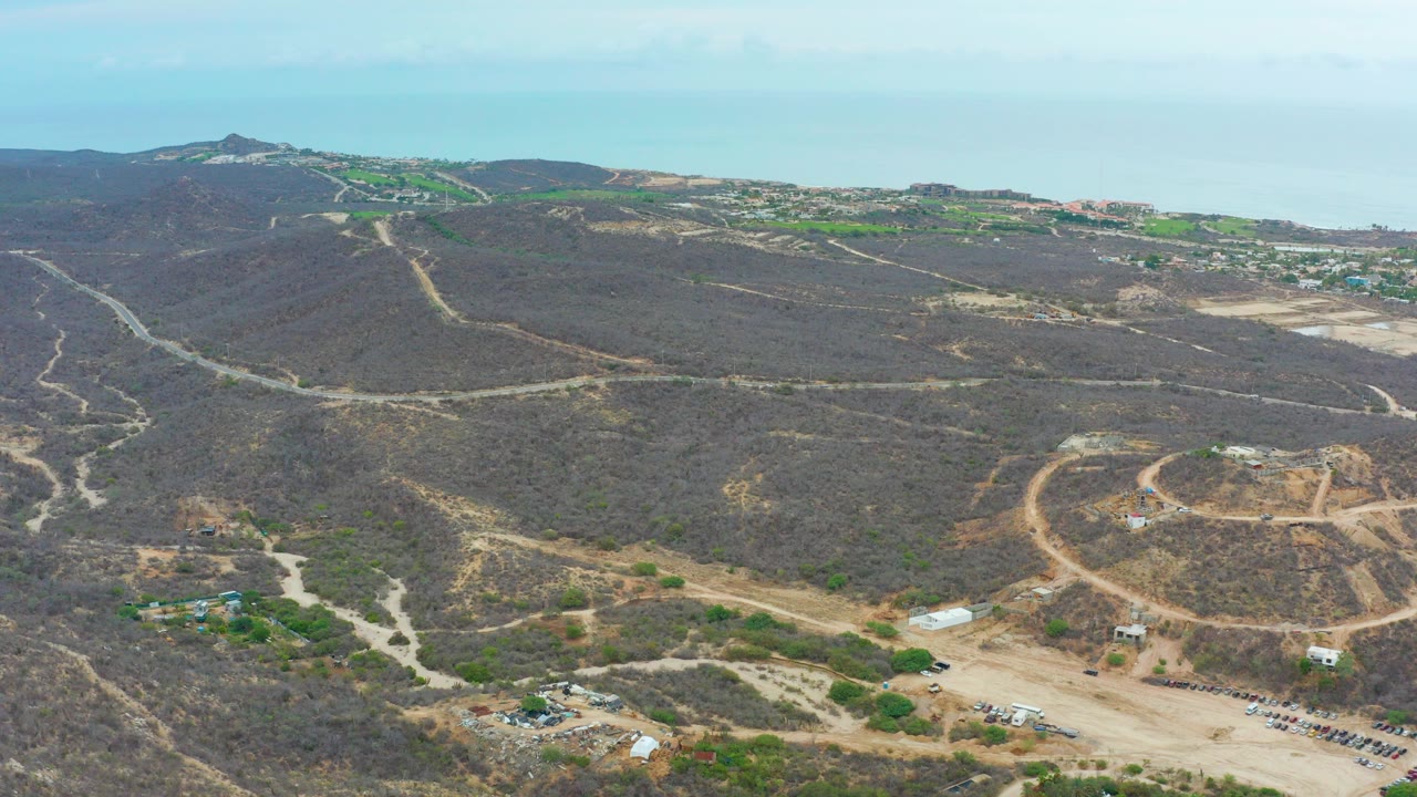 Wide aerial frame over La Playita Los Cabos showing dry hills crossed by pale dirt roads, scattered construction areas, a hazy ocean background, and no visible people.