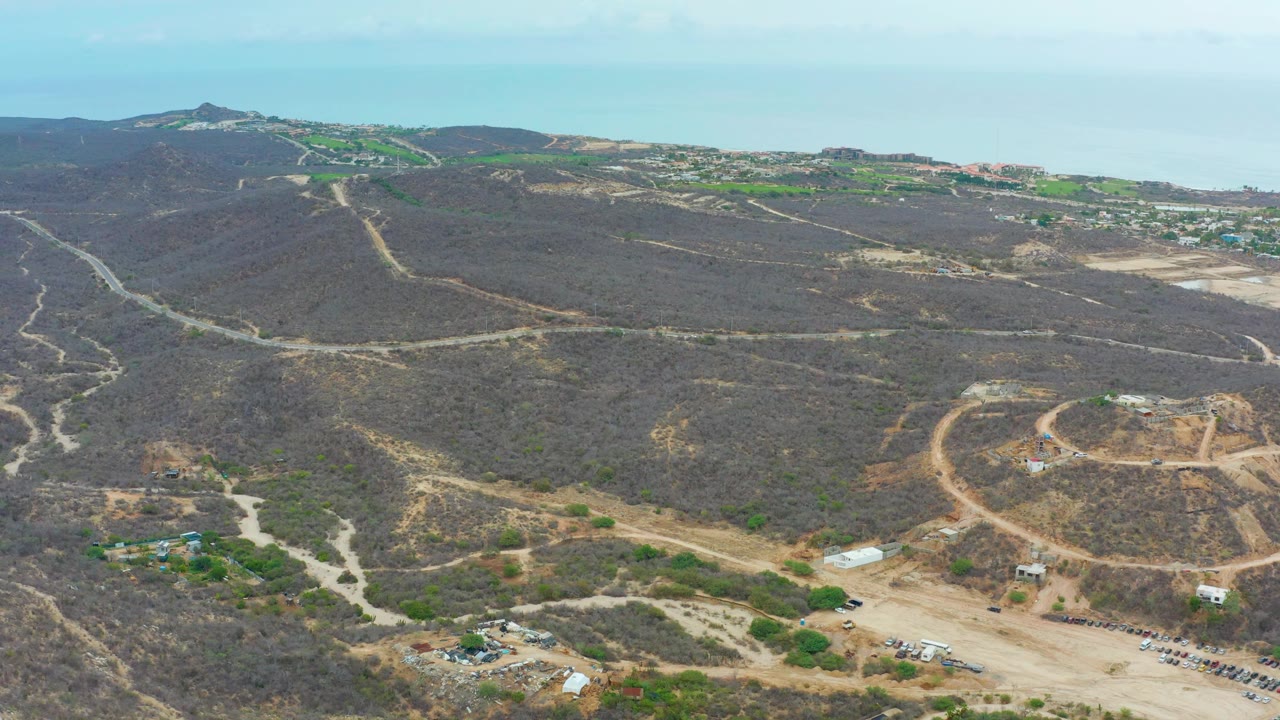Aerial landscape with dry ridges, dirt access roads, construction pads, and ocean beyond.