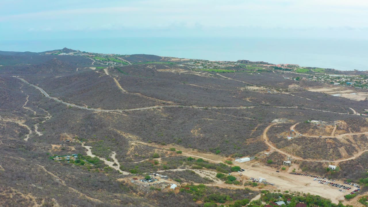 Aerial view over La Playita Los Cabos with scrub-covered hills, branching dirt tracks, a circular graded area, parked vehicles near the lower edge, distant settlement, and no visible people.