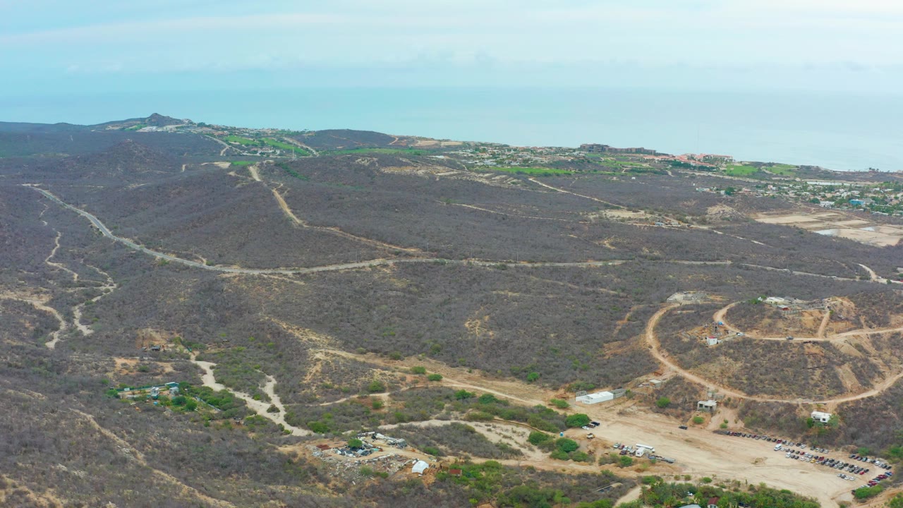 Mid-clip aerial over La Playita Los Cabos with dark brown hillside scrub, light dirt roads crossing the slopes, a graded lot and vehicles near the bottom, distant development, ocean horizon, and no visible people.