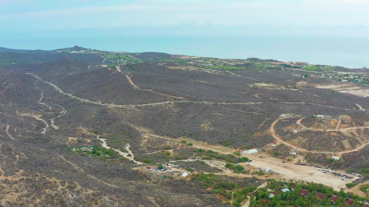 Aerial frame over La Playita Los Cabos showing brown hills, pale dirt roads, scattered green patches, distant water, and no visible people at the start of the clip.