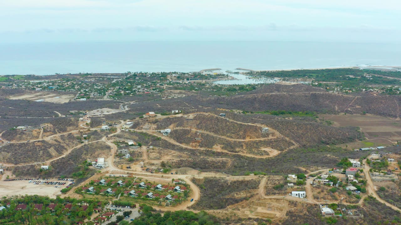 Aerial view over La Playita Los Cabos with dry foreground hills, winding dirt roads, scattered buildings, distant shoreline, pale ocean, and no visible people.