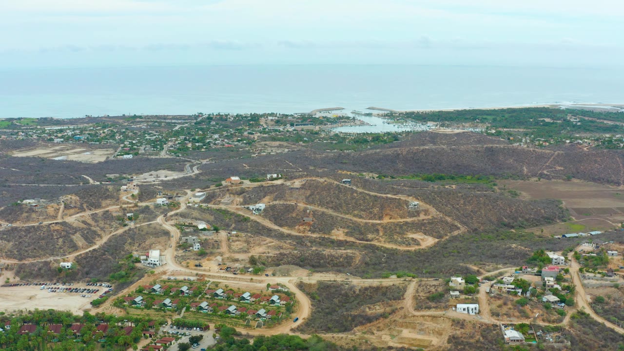 Elevated view in La Playita Los Cabos with curving dirt roads across dry hills, scattered houses or buildings below, a green coastal strip, ocean horizon, and no visible people.