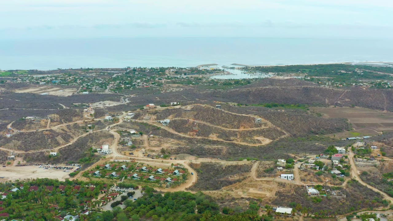 Wide aerial frame over La Playita Los Cabos, Mexico, showing dry hills, winding dirt roads, scattered homes, foreground greenery, distant ocean, and no visible people.
