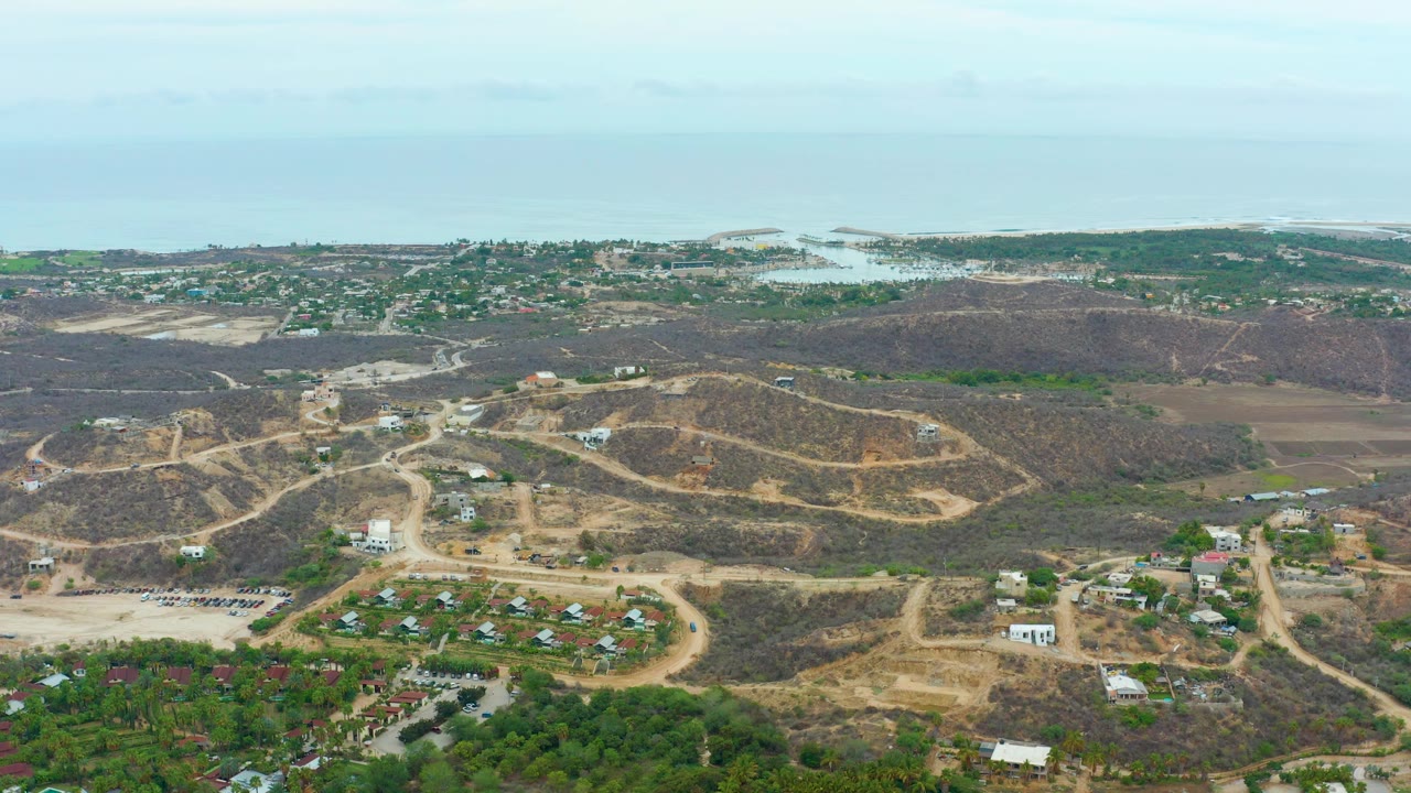 High drone frame over La Playita Los Cabos showing dirt tracks across brown hills, scattered residential buildings, green foreground lots, a distant coastal settlement, ocean water, and no visible people.