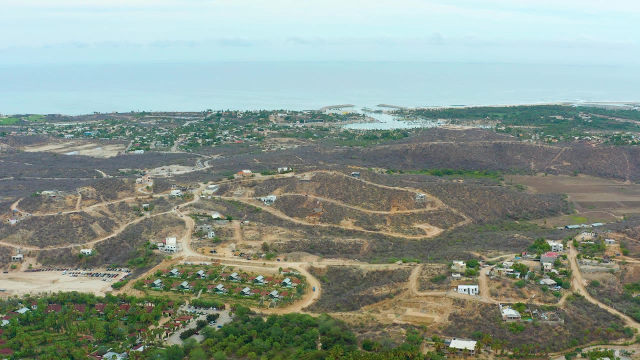 Aerial landscape of dry hills, homes, roads, greenery, and ocean horizon.